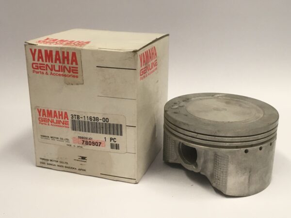 Una caja de piezas originales Yamaha está junto a un pistón de metal de 1,00 mm sobre una superficie lisa.