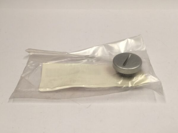Un pequeño botón redondo de plata con una ranura en la parte superior está sellado dentro de una bolsa de plástico transparente sobre una superficie blanca, listo para su próxima aventura como el Tapón pequeño culata.